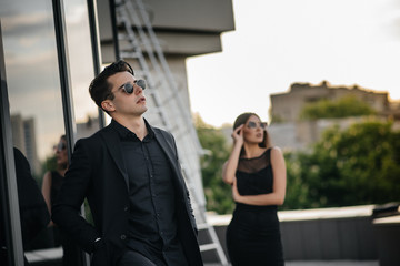 A beautiful, stylish pair of young people in black clothes and glasses stand against the background of an office building in the sunset. Fashion and style