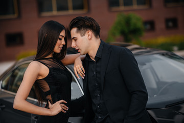 A young stylish couple in black stands near the car at sunset. Fashion and style