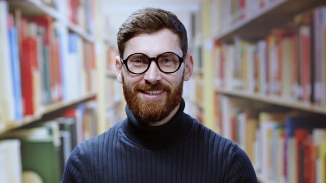 Close-up young bearded man wearing glasses looking shy smiling cheerful at camera standing between bookshelves working in the library.