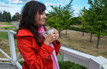 Pretty brunette girl in red coat holds cup in the autumn park and look into the distance.