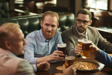 Group of colleagues sitting at the table drinking beer with peanuts and talking to each other in the pub