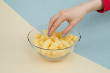 The hand reaches for the corn sticks in a glass bowl on a yellow-blue background. the concept of entertainment at home in quarantine coronavirus