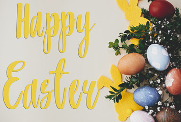 Happy Easter text. Easter greetings card. Stylish easter eggs, yellow bunny, spring flowers on yellow paper. Natural dyed easter eggs and rabbit decoration. Holiday card
