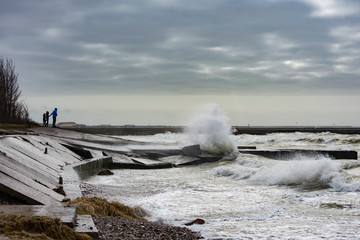 Man with children in front of big splash of water. Waves hitting concrete sea wall pier
