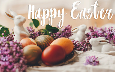 Happy Easter text. Easter greetings card. Stylish Easter eggs, white bunnies and lilac flowers on linen rural fabric. Natural dyed yellow easter eggs and spring flowers.  Holiday card