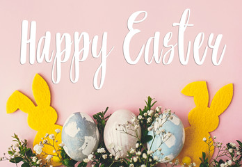 Fototapeta premium Happy Easter text. Easter greetings card. Stylish easter eggs, yellow bunny, spring flowers on pink paper flat lay. Natural dyed easter eggs and rabbit decoration. Holiday card