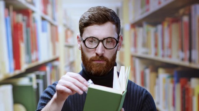 Happy bearded librarian or university student discovers pack of secret marijuana weed pack in a book smelling good smiling cheerfyl at camera staying in library.