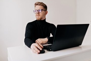 Stylish young european man in black pullover working on laptop in light office in sunny day
