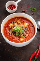 Spicy tomato cream soup with potatoes and green leaves in a white bowl on a rusty steel background