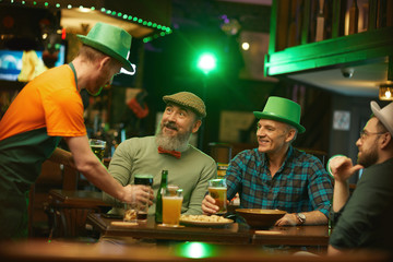 Group of men sitting at the table and talking to the waiter while he bringing glasses of beer for them in the pub