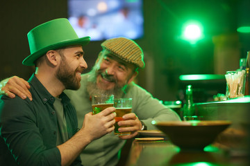 Two happy men in hats talking to each other laughing and drinking beer at the bar counter in pub