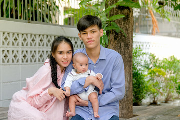 Asian mother is sitting with her husband who is holding their little baby in home garden with morning light and they look happy.