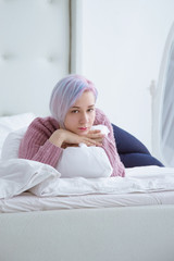 Beautiful young woman with blue hair lies on a bed in a bedroom dressed in lilac, purple sweater.