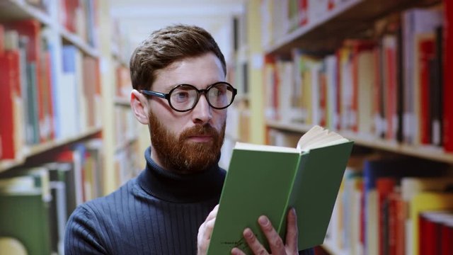 Portrait of tricky attractive man holding a book taking pictures on smartphone against rule naughty standing at bookshelves in the library.