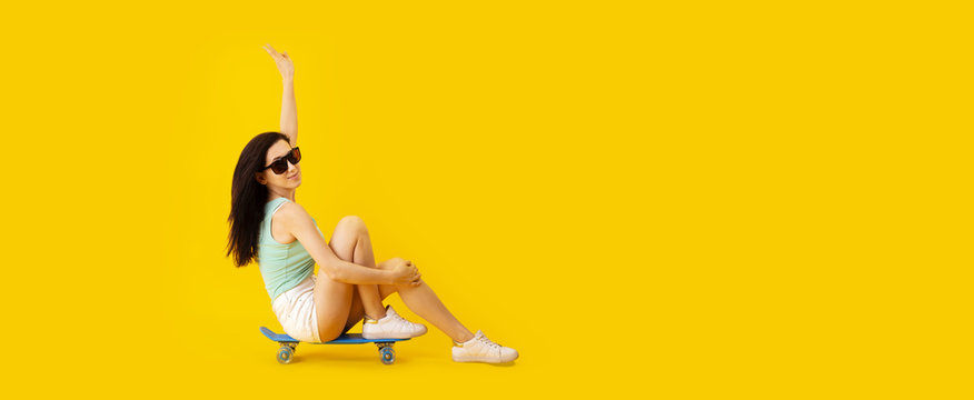 studio portrait of young girl in sunglasses sitting on skateboard over yellow trendy background, panoramic image