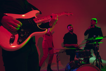 Obraz premium Young and joyful caucasian musicians performing on red studio background in neon light. Concept of music, hobby, festival. Colorful portrait of modern artist. Attented and inspired. Art, cover band.