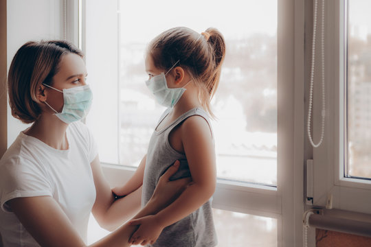 Family Mom and Daughter in Medical Mask. Young Woman and Child little girl sitting by the Window in Protective Masks against the Virus. Little girl in Hospital.      
