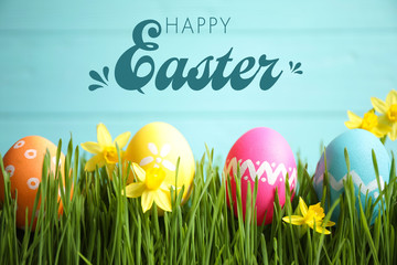 Colorful eggs and narcissus flowers in green grass and text Happy Easter against light blue background