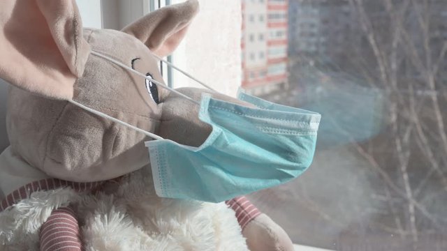 A plush toy rat in a medical protective mask sits on a windowsill and looks out into the street. Sadness and loneliness during quarantine at home. Symbol of 2020.