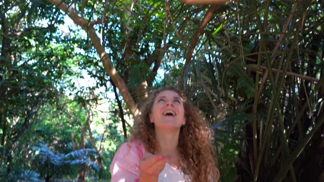 A curly-haired blond model in pink clothes feeds and plays with a squirrel monkey on a trip to a tropical island in Japan. A bright smiling emotional girl in a natural zoo gives a cute saigiri monkey 