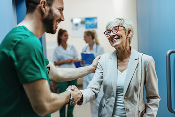 Happy senior woman shaking hands with a surgeon in a hospital hallway.