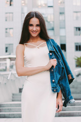 Portrait of gorgeous young cute trendy hipster girl in stylish denim jacket and dress posing and smiling on city street. Beautiful cheerful fashionable woman walking outdoors. Modern city fashion