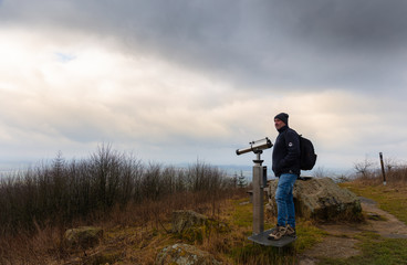 A hiker with a backpack stands on the highest mountain in East Westphalia with the name Köterberg. Next to the man are silver binoculars with a coin slot.