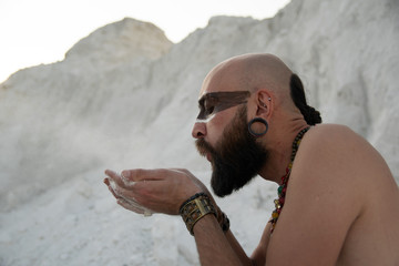 young man with primal makeup and beads spells with powder in hands on white rocks