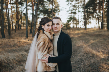 Stylish groom in a black cardigan and a beautiful bride in an expensive dress are hugging in the forest on the nature. Wedding portrait of smiling newlyweds. Photography, concept.