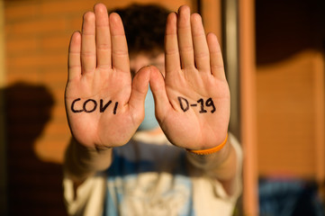 Close-up image of the palms of the hands with the inscription COVID-19 of a Caucasian boy with curly brown hair wearing a blue mask. Conceptual image of danger of contagion. Selective focus on palms.
