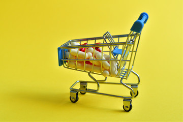 Shopping trolley cart with assorted medicine pills on yellow background. Creative idea for drugstore, online pharmacy, pharmaceutical company business concept.