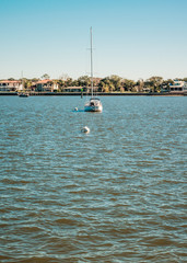 Obraz premium Small Sailboat Lying At Anchor with Houses Along the Coast Behind