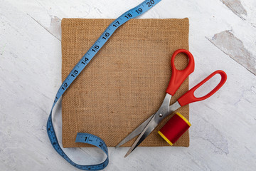 Brown burlap square craft canvas with measuring tape spool of red yarn thread and scissors 