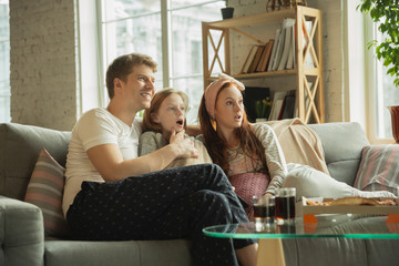 Family spending nice time together at home, looks happy and cheerful. Mom, dad and daughter having fun, eating pizza, watching sport match or TV. Togetherness, home comfort, love, relations concept.