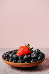 rustic handmade plate full of blueberries with a strawberry on top ready to eat placed on a cement textured background and pink background