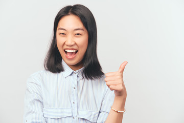 Girl holds finger up, indicating that great idea. Beautiful young Asian appearance with black hair and brown eyes dressed in striped office shirt stands isolated white background in Studio