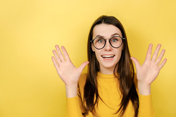 Portrait of impressed beautiful young woman in spectacles spreads palms with excitement, smiles broadly, feels amused, wears sweater, poses over yellow background with copy space