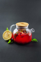 glass transparent teapot with strawberry tea and cut orange and mint on a black background