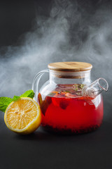 glass transparent teapot with strawberry tea and mint on a black background with smoke