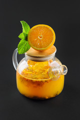 glass transparent teapot with sea buckthorn tea and cut orange and mint on a black background