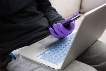 Selective focus on student hand with medical glove holding cell phone one hand and working with a computer typing on a laptop at home. Quararntin remote work and study during coronavirus pandemic