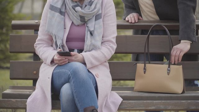 Male Caucasian hand stealing wallet from purse of unrecognizable woman absorbed with smartphone. Adult lady losing money in park as messaging. Crime, pickpocketing, device addiction.