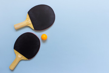 Flat lay with two black table tennis rackets and orange ball on blue background. Photo with copy blank space.