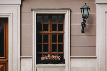 Old brown wooden window with rectangular frames for glass, a flower pot with flowers, a door, a wall lamp and a house gray facade.