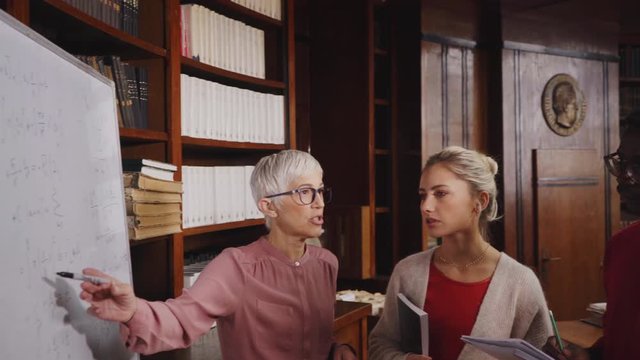 Focused university students talking with teacher in library. College professor with multiethnic class studying in library. Group of four focused clever students in conversation with senior teacher.