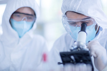 A male scientist wearing a mask and looking through the microscope in the laboratory, Concept, The scientists wear white protective clothing is experimenting with drug anti- Coronavirus, Covid19