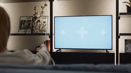 Over shoulder close up view of young woman changing channels with a TV remote control on a green white chroma key screen mock-up with track points sitting on sofa relaxing in cozy home warm lights