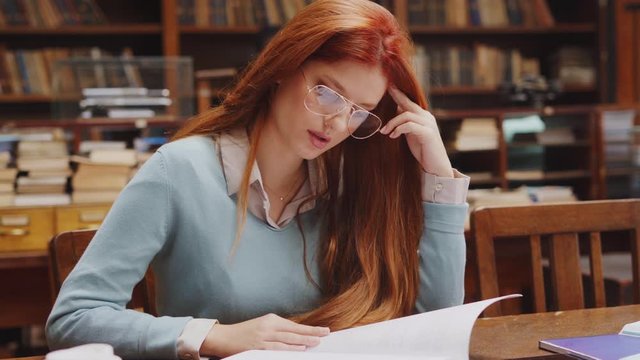 Woman with spectacles sitting in library and reading. Portrait of university student studying with concentration in old library. Girl with red hair sitting and doing assignment in college library.