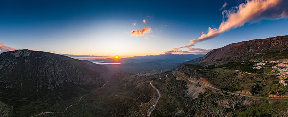 Fototapeta premium Aerial view of Delphi, Greece, the Gulf of Corinth, orange color of clouds, mountainside with layered hills beyond with rooftops in foreground