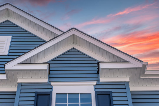 Triangle shape decorative gable with colonial white soffit and fascia on a blue horizontal vinyl siding modern American estate home with colorful dramatic stunning orange sunset sky